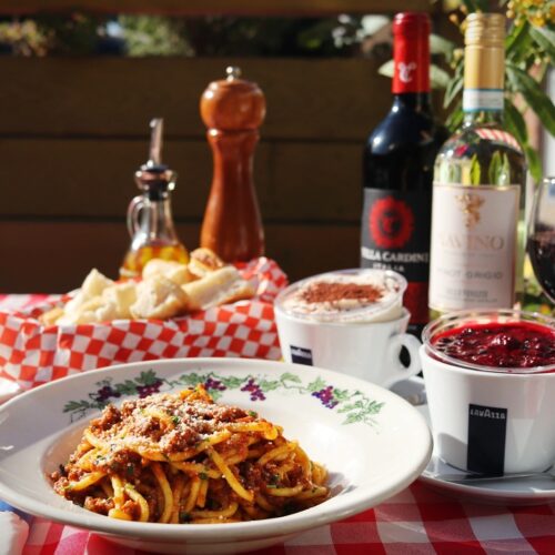 Spaghetti Ragú Bolognese with bottles of red/white wine, basket of bread, pepper mill, olive oil and dessert on table.